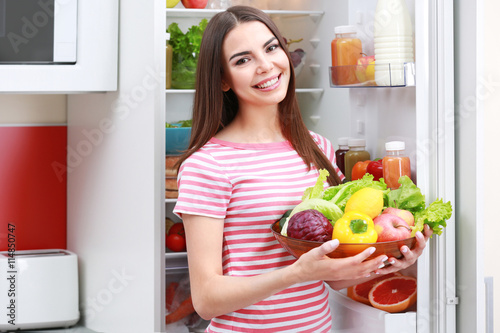 Young woman with fruits and vegetables beside fridge in kitchen