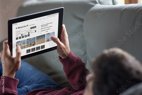 Young man using his tablet lying on the couch, fictional travel agency web