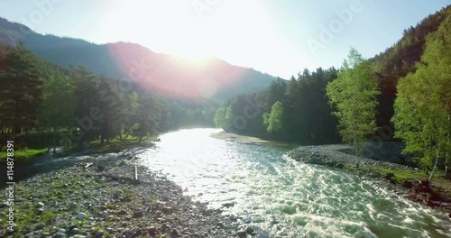4k (UHD) aerial view. Low flight over fresh cold mountain river at sunny summer morning. Green trees and sun rays.