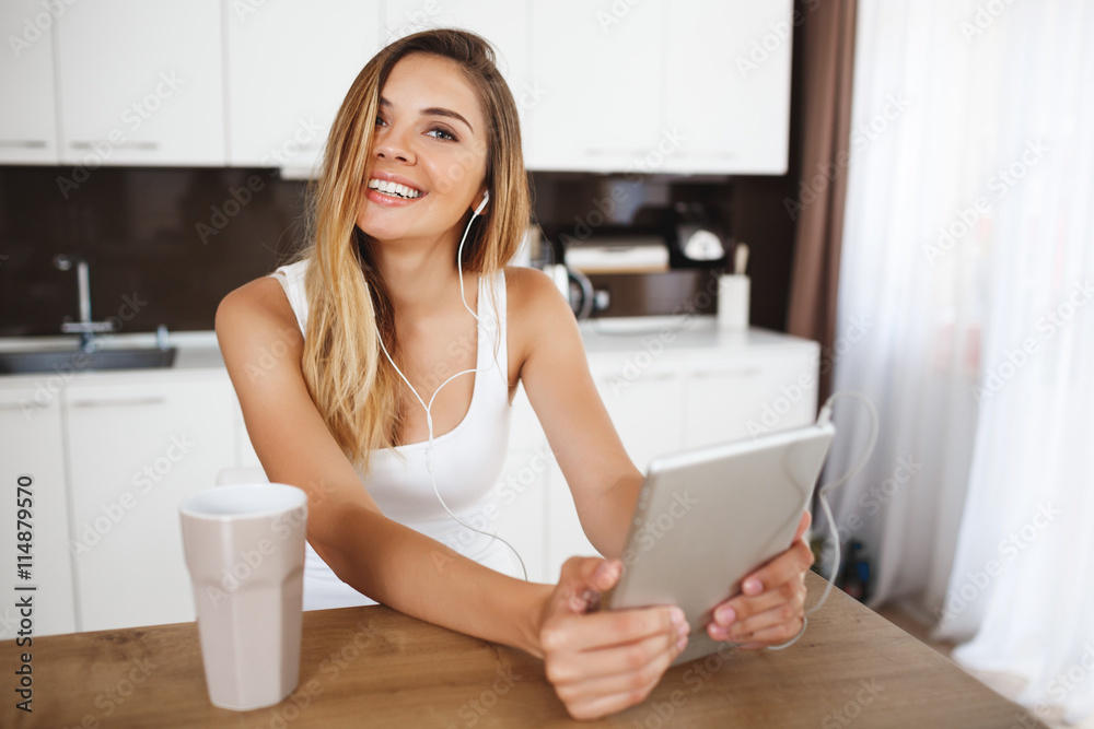 Attractive smiling young girl sitting at dinner table and holding tablet. Attractive smiling young girl sitting at dinner table and holding tablet.