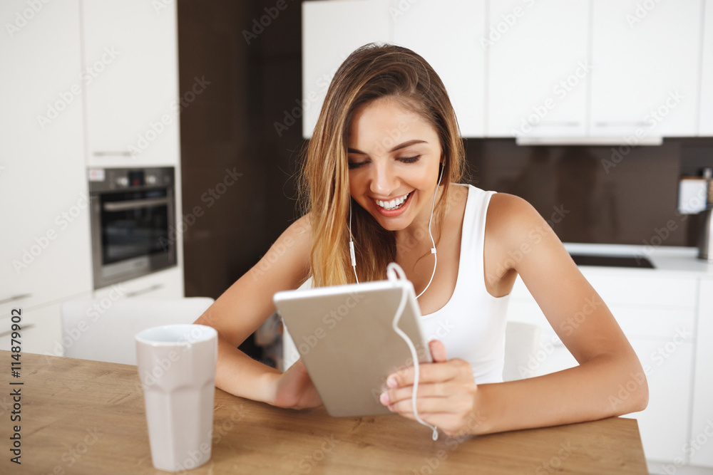 Attractive smiling young girl sitting at dinner table and listen to music and cheking something in tablet Attractive smiling young girl sitting at dinner table and listen to music and cheking something in tablet