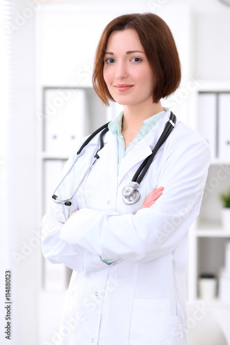 Young brunette female doctor standing with arms crossed and smiling at hospital. Health care, insurance and help concept. Physician ready to examine patient