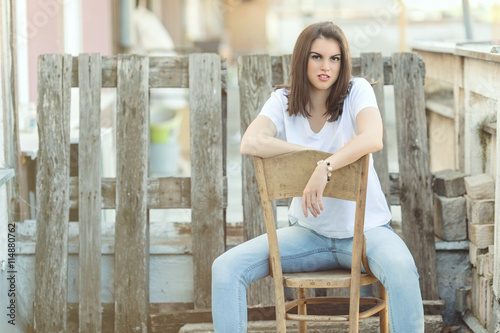 Portrait of a beautiful young woman sitting on a chair in front of the rustic wooden fence on a rooftop terrace and enjoying sunny summer day.