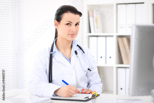 Young brunette female doctor sitting  with clipboard near window in  hospital and filling up medical history form. © rogerphoto