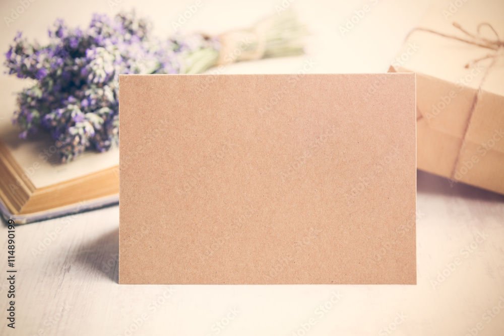 Empty greeting kraft card in front of a lavender bouquet, wrapped gift and old book over a white wood background. Vintage style.