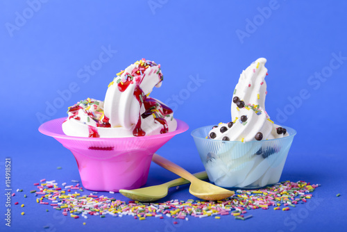two frozen yogurt with black cherry topping and rainbow sprinkles and chocolate candy on blue background with yellow and green spoons and rainbow sprinkles