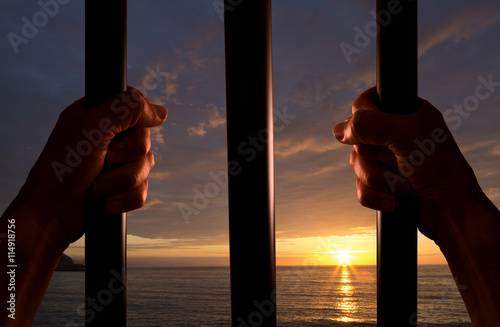 Photo Hands of the prisoner on a steel lattice close up with the sunset in the backgro
