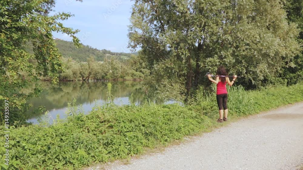 Beautiful young woman doing back stretching after nordic walking session on the riverside during a sunny day in summer. Girl doing outdoor sport. Take care of yourself. Health, wellness and beauty Beautiful young woman doing back stretching after nordic walking session on the riverside during a sunny day in summer. Girl doing outdoor sport. Take care of yourself. Health, wellness and beauty