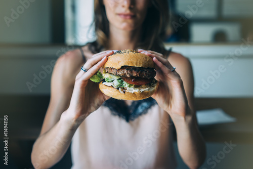 Young girl holding in female hands fast food burger, american unhealthy calories meal on blue background, mockup with copy space for text message, hungry human with grilled hamburger