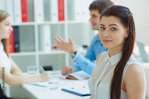 Beautiful smiling business woman loking straight to the camera while couple young business partners discussing ideas for a startup in background. Business and partnership concept.