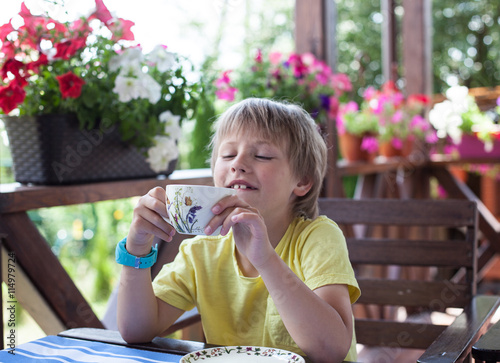 Smiling boy with smart watch sitting in wooden armchair and drinking tea on a terrace  