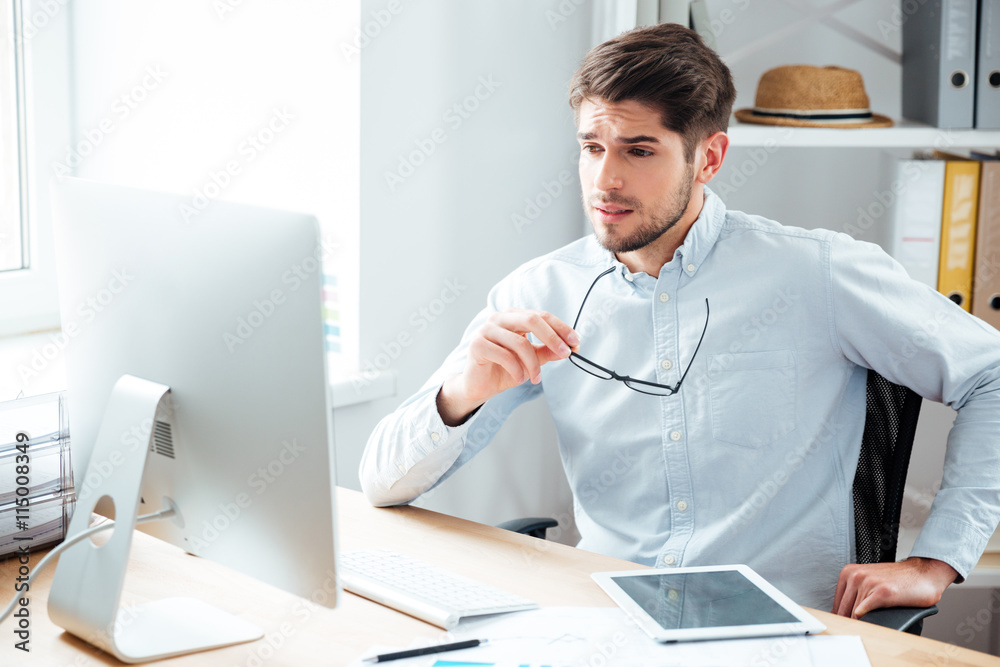 Fototapeta premium Handsome businessman holding eyeglasses and looking at computer monitor