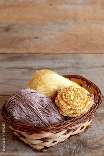 Crocheted yellow rose with brown leaves and two skeins of cotton yarn in a basket. Beautiful knitted flower adorned with beads. Stylish crochet crafts. Old wooden background with empty space for text