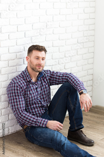 Handsome young man smiling while sitting on the floor and leaning at the wall