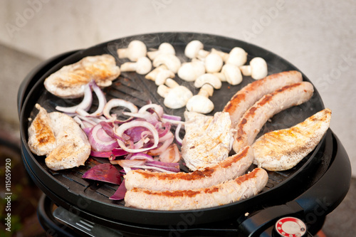 Selection of meat grilling on a electric portable barbecue with sausages, champignons and onion. Bbq. BBQ season.