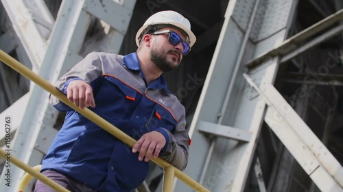 Industrial worker in protective helmet and sunglasses stands in front of metal structure. photo