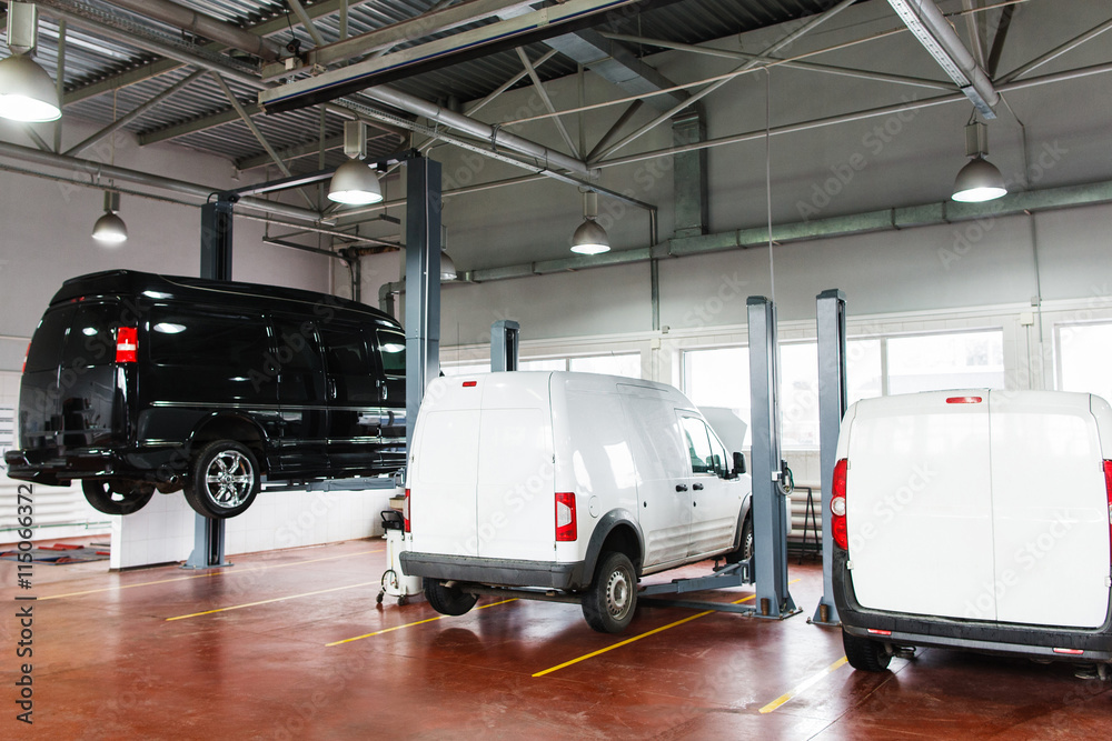 SUV big service station, black minibus raised up on lift. Front view on line of three cars in garage on service maintenance, two white pickups stay in stables, black minibus lifted up SUV big service station, black minibus raised up on lift. Front view on line of three cars in garage on service maintenance, two white pickups stay in stables, black minibus lifted up