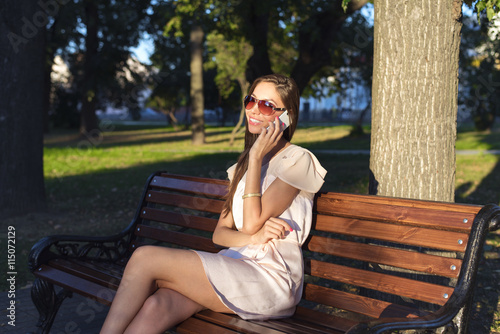 Beautiful emotional brunette girl laughing on the phone in the park summer glasses pink dress, enjoy your holiday in the city. Fashion style of life, a bright day. The tree the bench.