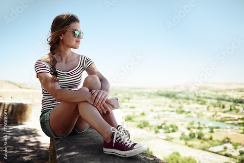 Pretty woman in casual outfit sitting on stone looking at beautiful scene of countryside. Young teenage girl meditating or relaxing alone in silence. 