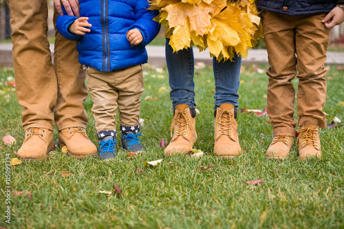 Closeup of legs of a family standing on green grass and holding bunches of yellow marple leaves  outdoors on autumn day. Couple and children holding hands and enjoying walk in park. Lifestyle concept