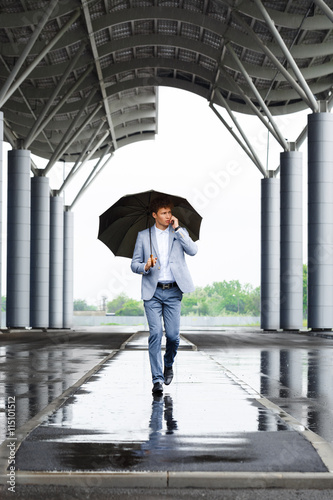 Picture of handsome young redhaired businessman holding umbrella and talking on phone walking in street © Cookie Studio