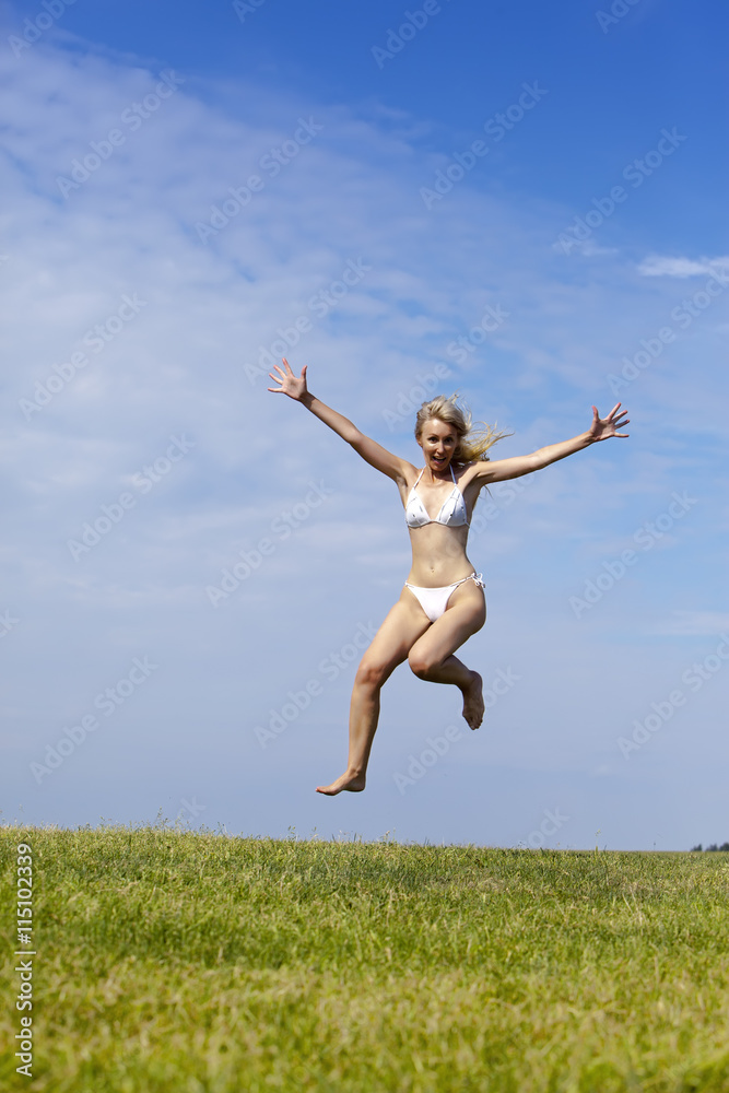 The happy woman in white bikini  jumps in a summer green field against the blue sky.