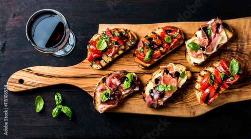 Brushetta set and glass of red wine. Small sandwiches with prosciutto, tomatoes, parmesan cheese, fresh basil, balsamic creme on rustic wooden board