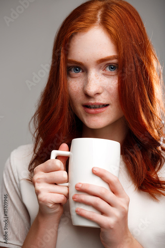 Portrait of beautiful ginger girl holding cup over gray background.