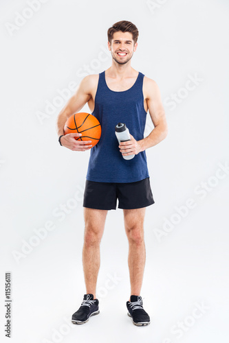 Obraz na plátně Smiling young sportsman holding basket ball and water bottle