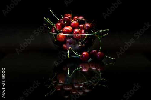 Transparent cup with red ripe cherries with reflection on black background.