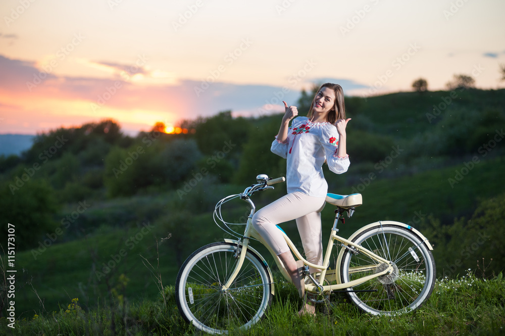 Close-up of smiling female with retro bicycle standing at hill and showing thumb up gesture of good class against beautiful sunset with a blurred background of greenery Close-up of smiling female with retro bicycle standing at hill and showing thumb up gesture of good class against beautiful sunset with a blurred background of greenery