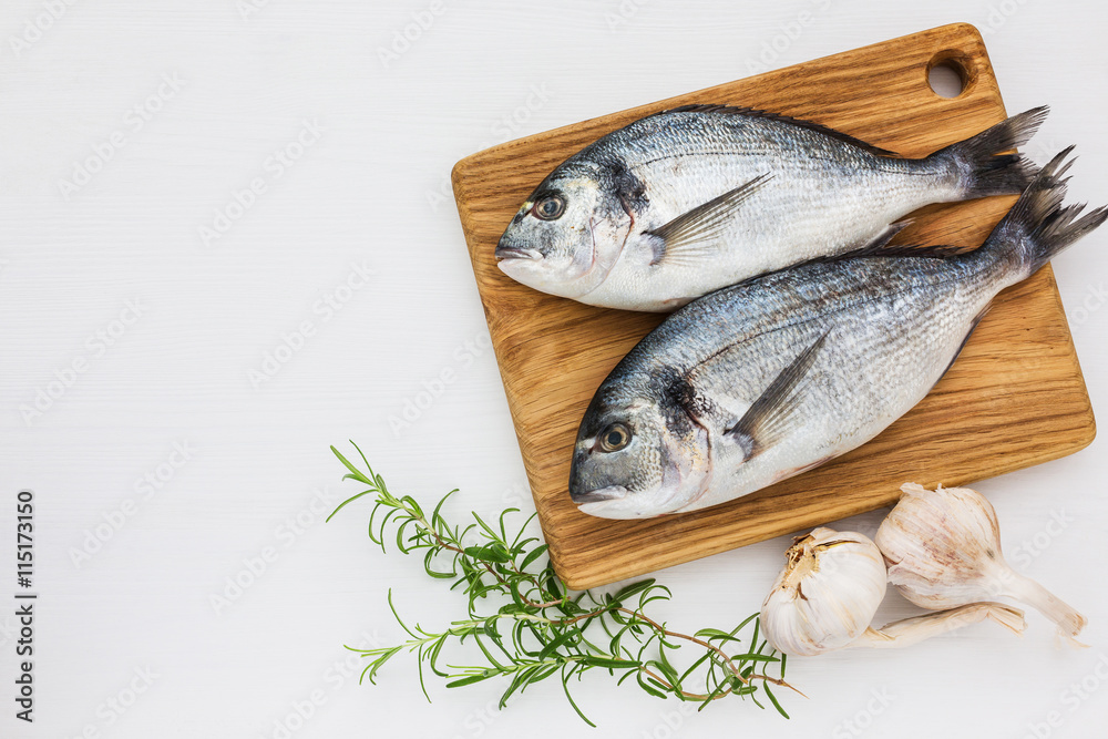 Fresh dorado fish on wooden cutting board with garlic and rosemary on white table. Top view, copy space. Fresh dorado fish on wooden cutting board with garlic and rosemary on white table. Top view, copy space.