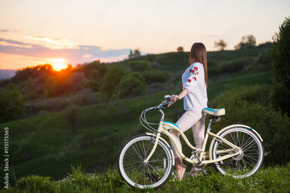 Woman in Ukrainian embroidery with bicycle standing at hill and looks at beautiful sunset with a blurred background of greenery