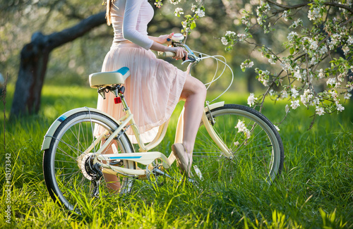 Close-up of graceful bend leg of pretty girl in white dress sitting on bicycle in the fresh greenery near blossoming tree in the ray of sunshine