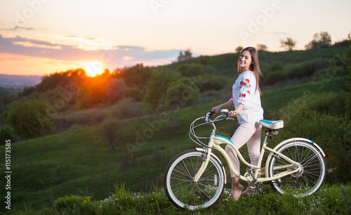 Happy girl with retro bike standing at hill and looks to the camera, with a blurred background of greenery and beautiful sunset