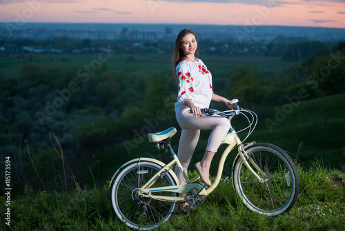 Happy young woman holding retro bicycle at hill on a blurred background of greenery and small town at sunset