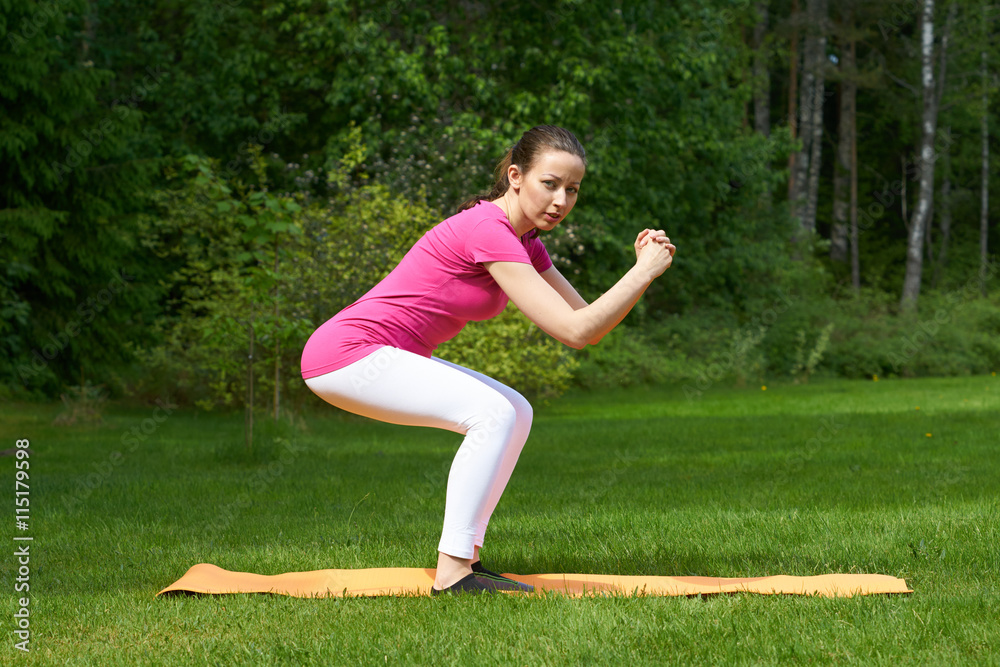 Attractive brown haired woman in pink t-shirt, white leggings, black socks with green stripers training buttocks and legs muscles in squat on orange mat in outdoor on grass in nature. Attractive brown haired woman in pink t-shirt, white leggings, black socks with green stripers training buttocks and legs muscles in squat on orange mat in outdoor on grass in nature.