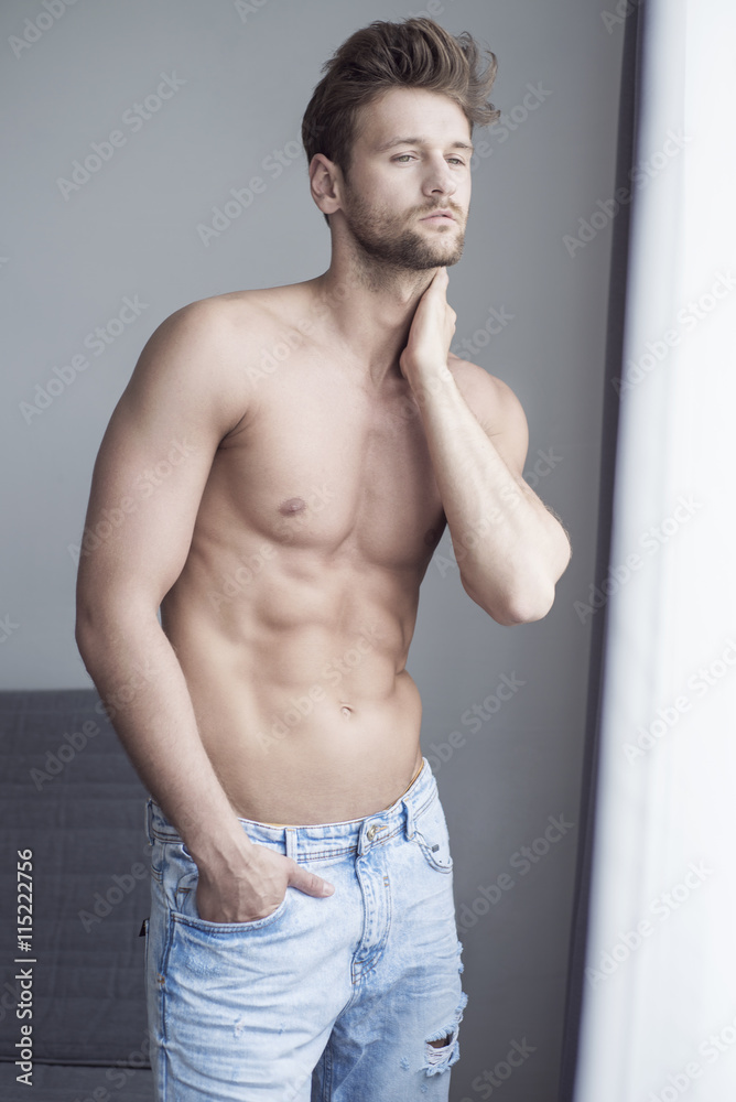 Fototapeta premium Handsome young man well-built stands at the window in the apartment