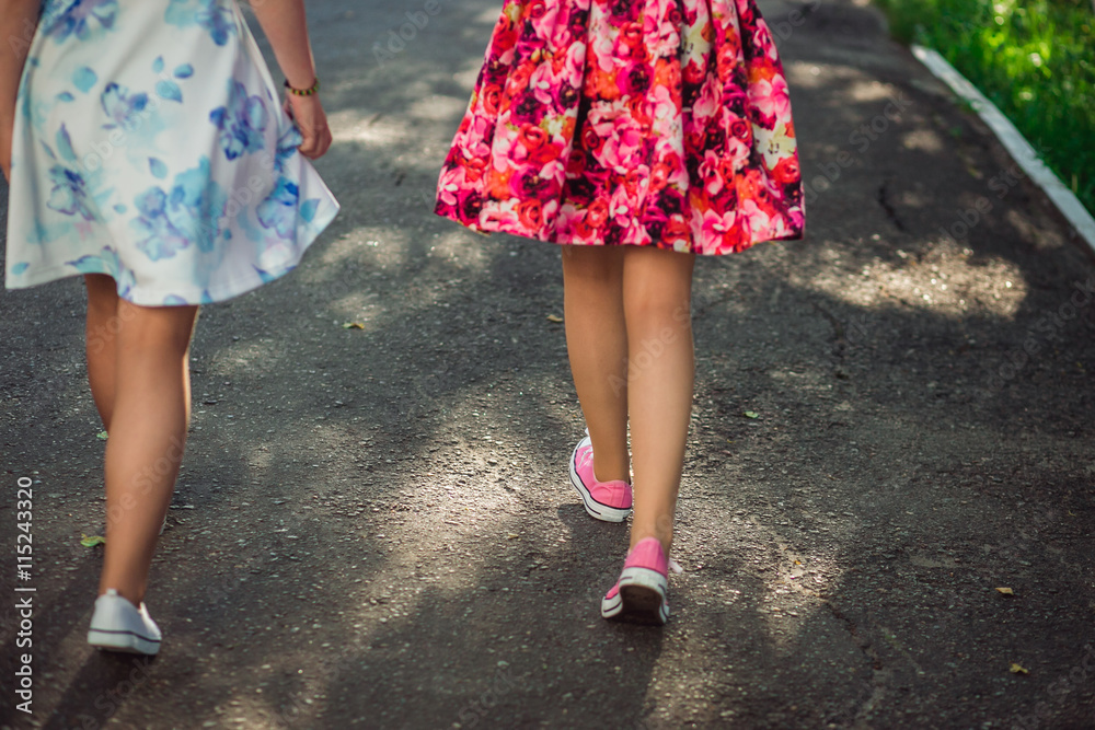Happy Teenage girls in light dresses walking on sun light, outdoors. Unrecognizable girlfriends having fun together. Closeup back view of legs.