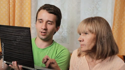 Adult male teaches old woman working for a laptop. Mother and son sitting on a sofa at home