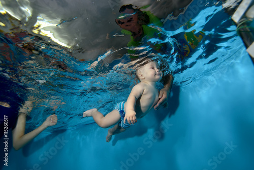 Happy baby trains and swims underwater in the pool on a blue background, and the dad catches him. Bottom view. Horizontal orientation