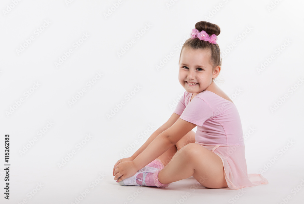 Little happy girl gymnast sitting on the floor holding his leg. The girl smiles. Light background and space for text