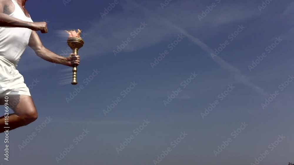 Torchbearer athlete in white running with sport torch in glowing slow motion across sunny blue sky