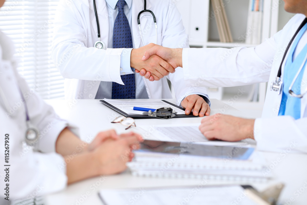 Two doctors shaking hands to each other sitting at the table in hospital office Two doctors shaking hands to each other sitting at the table in hospital office