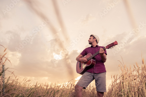 Young retro styled man playing acoustic guitar in wheat field. Sun and clouds in the background. Music, art and lifestyle concepts. 