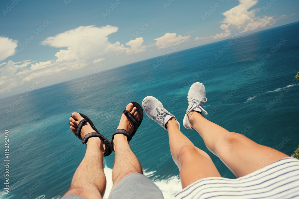 Φωτογραφία Legs of man in sandals and woman in sport shoes sitting above the ocean