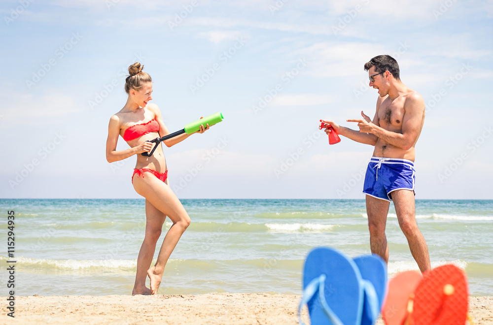 Playful young couple having fun on the beach - Sexy girl with water gun  ready for summer holiday games - Concept of childish joyful moment and  popular teens activities at summertime Stock