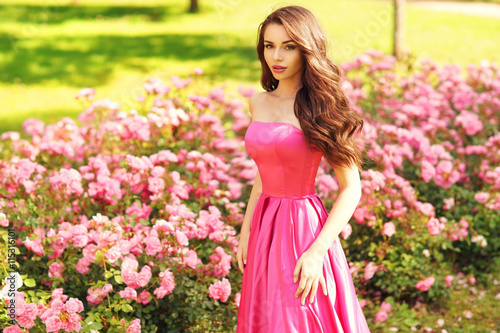 Young beautiful pretty woman posing in long evening luxury dress against bushes with pink roses on a sunny summer day. Vogue style fashion sensual portrait