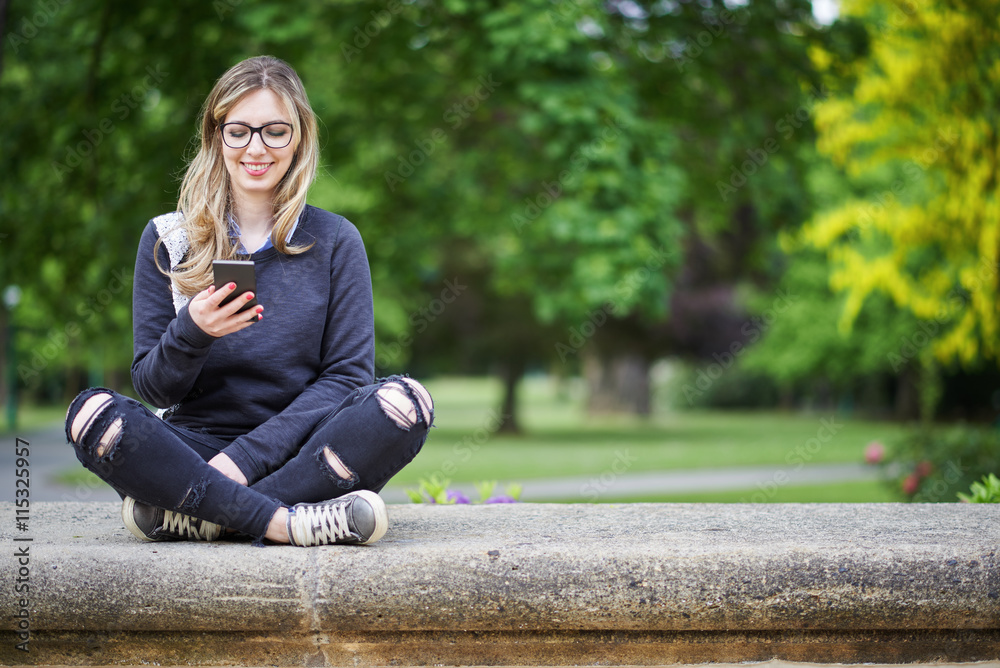 Young woman sitting on the wall in the park, writing a message on her mobile phone, park in the background