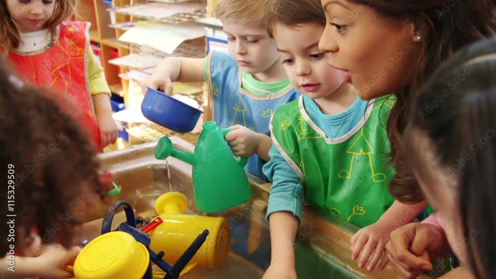 Nursery children playing at the water table in their classroom. The teacher is talking to them.